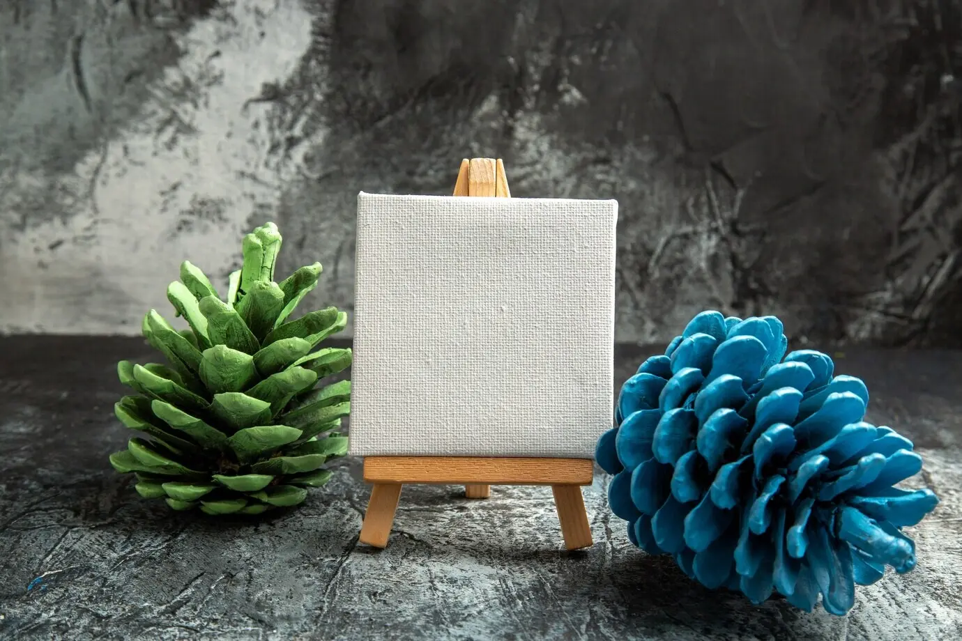 Vorderansicht einer kleinen weißen Leinwand auf einer Holzstaffelei mit farbigen Tannenzapfen vor dunklem Hintergrund.